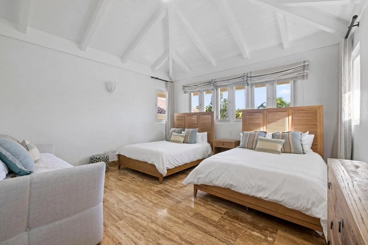 Habitación con dos camas de madera, un sillón y varias ventanas con cortinas, piso de madera clear y paredes blancas.