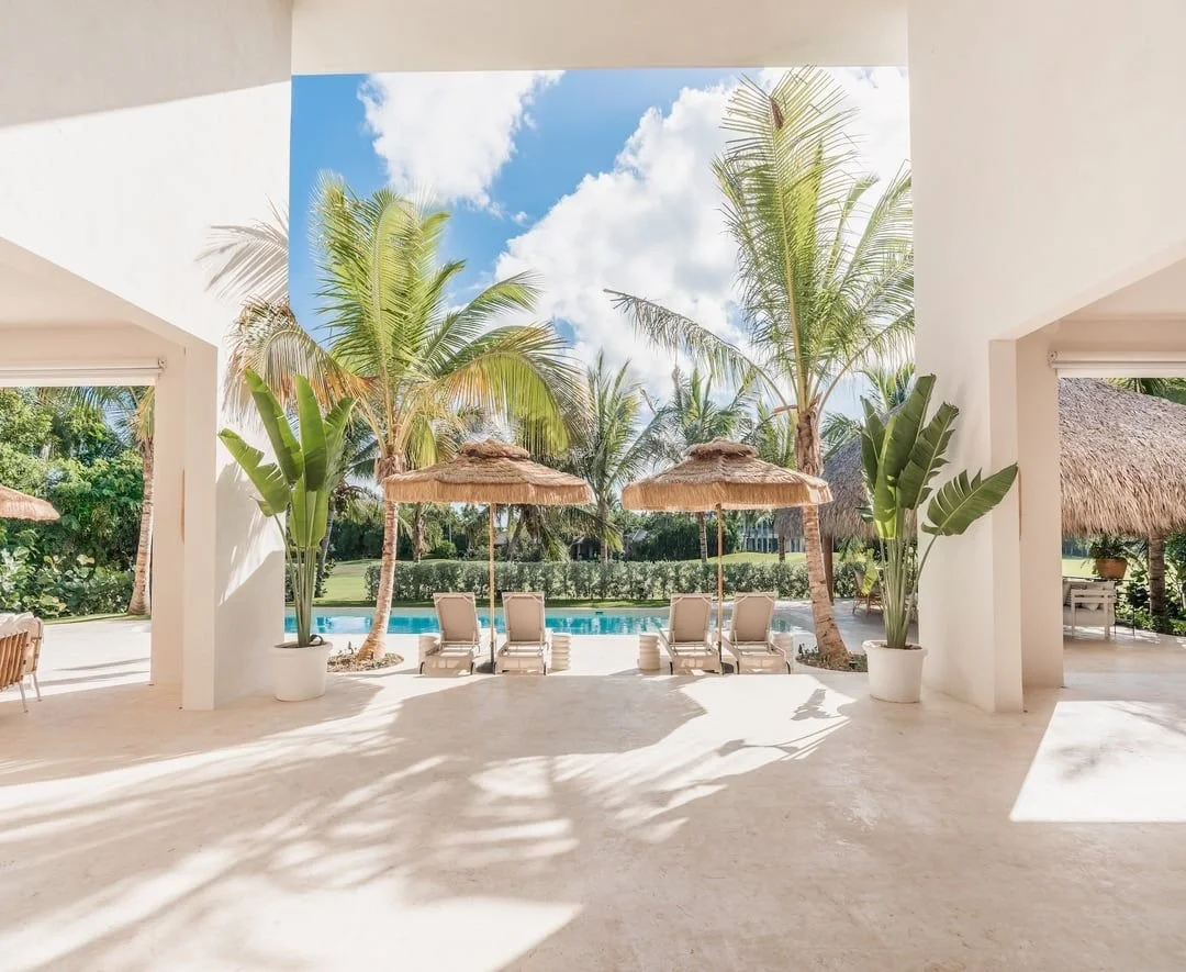 Vista de una piscina con tumbonas y sombrillas de paja, rodeada de palmeras y plantas verdes, bajo un cielo azul con nubes.