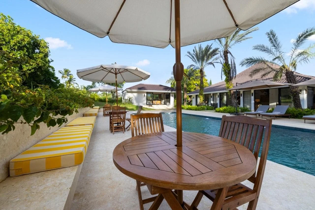 Área de alberca con mesas, sillas y sombrillas en un entorno tropical con casas y palmeras