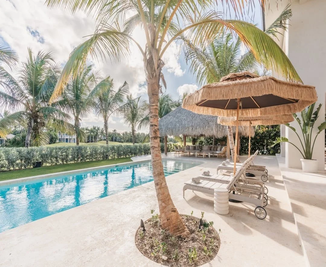 Piscina en un área de descanso con sombrillas de paja, tumbonas y palmeras.