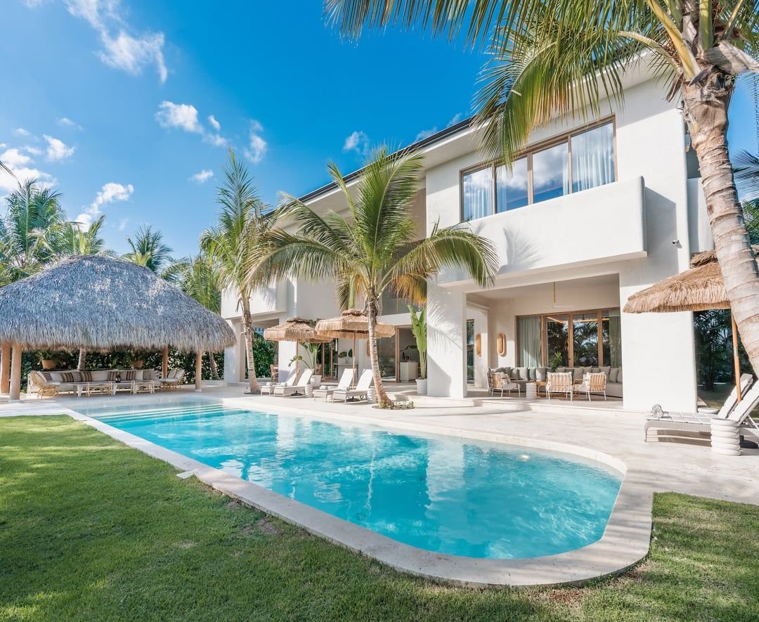 Casa moderna de dos pisos con piscina y área de descanso con sombrillas de paja y sillones, rodeada de palmeras en un día soleado.