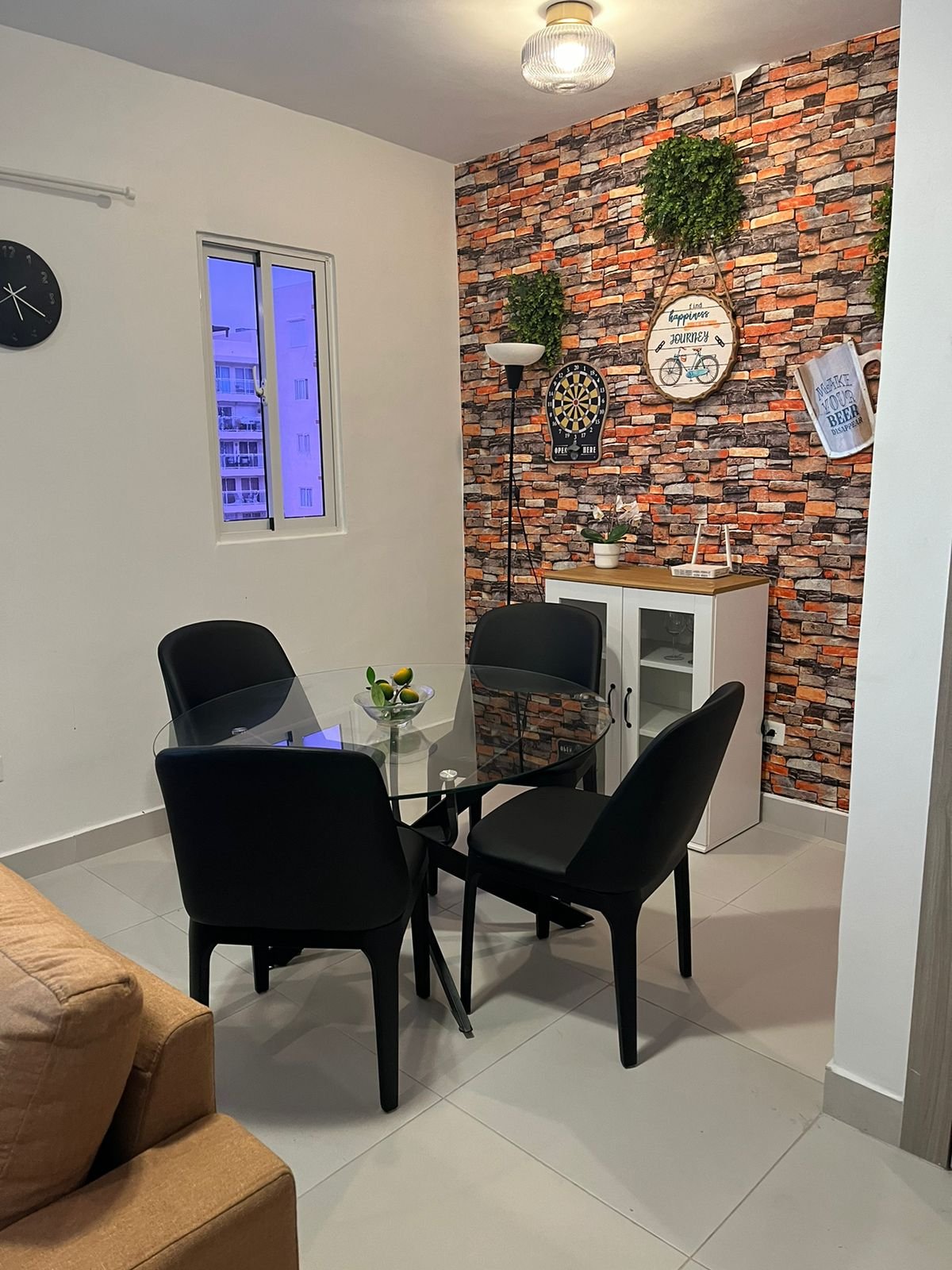 Comedor con mesa de vidrio y cuatro sillas negras, pared de ladrillos decorativos con plantas, reloj y arte en la pared, ventana y lámpara en el techo.