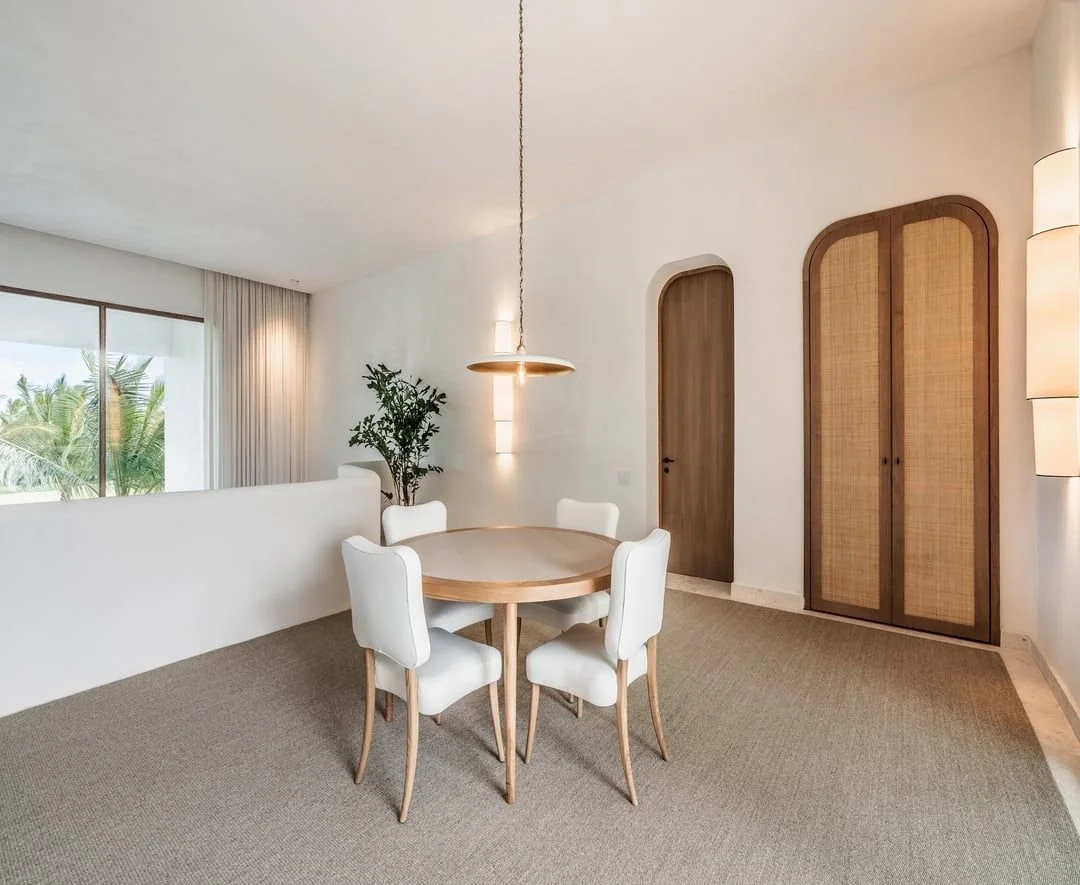 Sala de comedor con mesa redonda, seis sillas blancas, lámpara colgante de diseño minimalista, pared blanca, puertas de madera, ventana grande con cortinas, planta en esquina y paredes con iluminación indirecta.