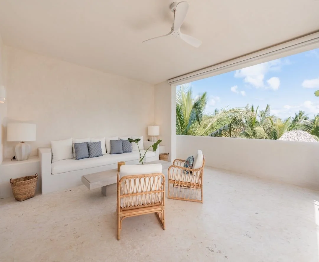 Salón con paredes blancas, sofá blanco con cojines grises, dos lámparas de mesa, una planta en una mesa y sillas de mimbre, vista a palmeras y cielo azul.