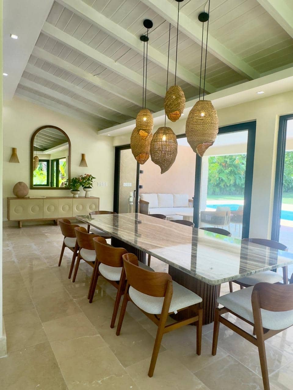 Interior de comedor con mesa de mármol y sillas de madera con cojines blancos, lámparas de fibra natural colgantes, espejo en la pared, ventanales con vista a jardín y piscina.