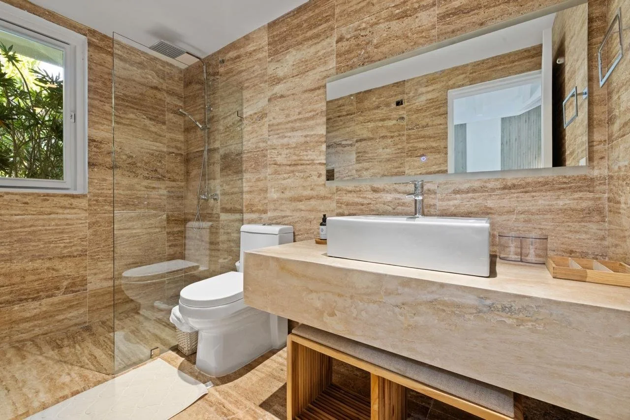 Baño con paredes y piso de piedra beige, lavabo rectangular blanco sobre encimera de piedra, gran espejo rectangular, inodoro blanco, ducha con mampara de vidrio, ventana con vista a plantas verdes, accesorios de baño de madera y vidrio.