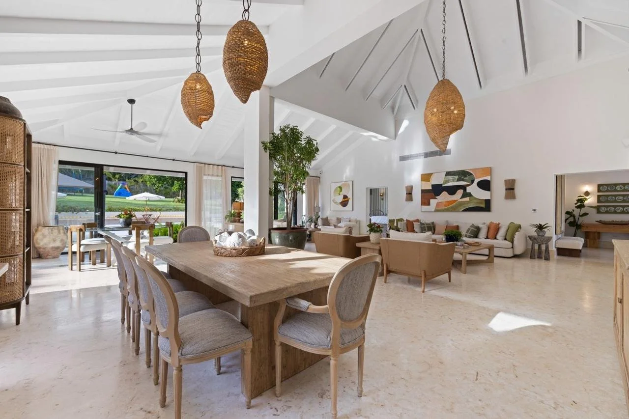 Interior de sala de estilo moderno y minimalista con mesa de madera, sillas, sofás y decoración en tonos neutros y naturales. Ventanas grandes con vista al exterior y lámparas colgantes de ratán.