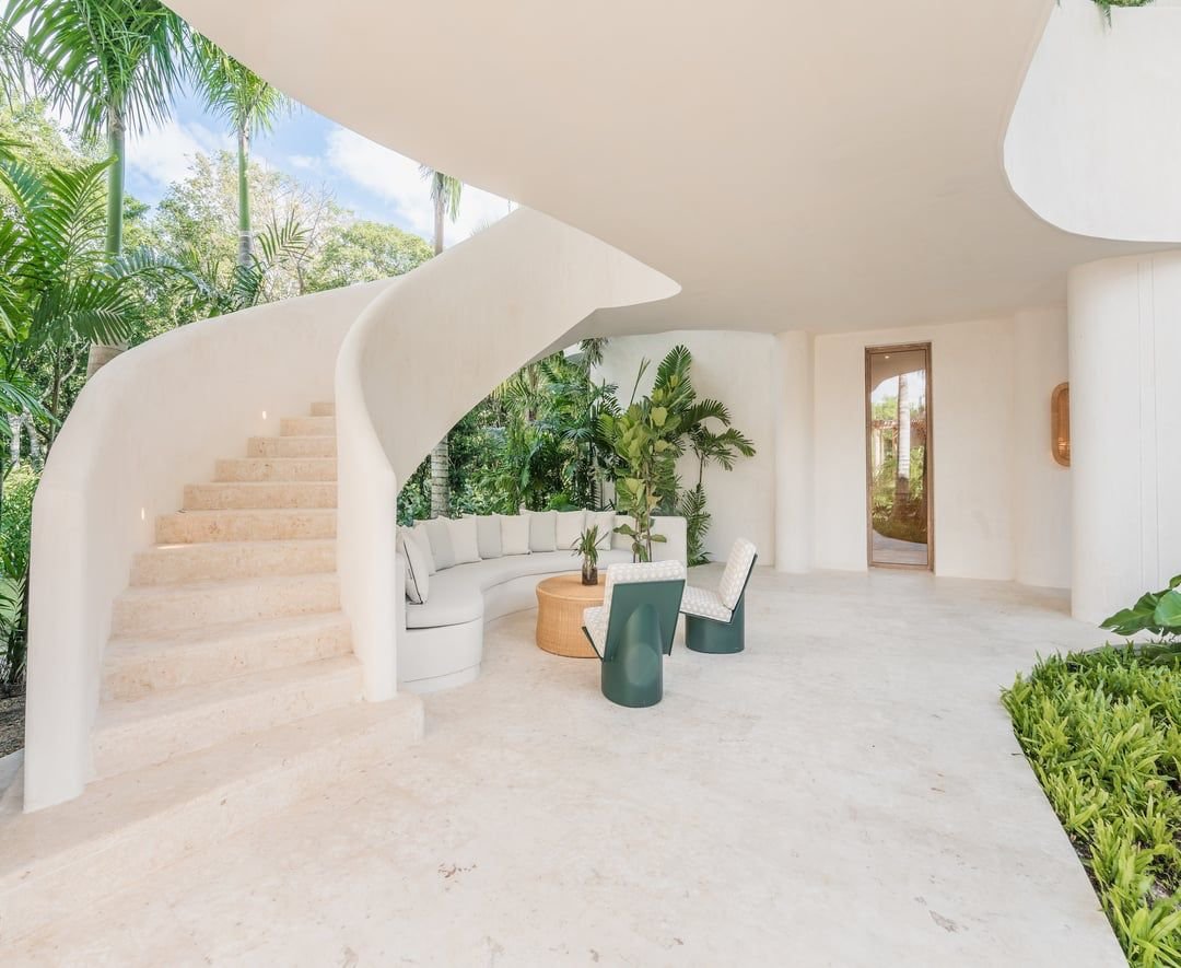 Espacio interior con paredes blancas, sofá en forma de esquina, mesa de centro de palma, sillas blancas, muchas plantas verdes, escaleras curvas blancas, y un fondo con vista a la naturaleza tropical.