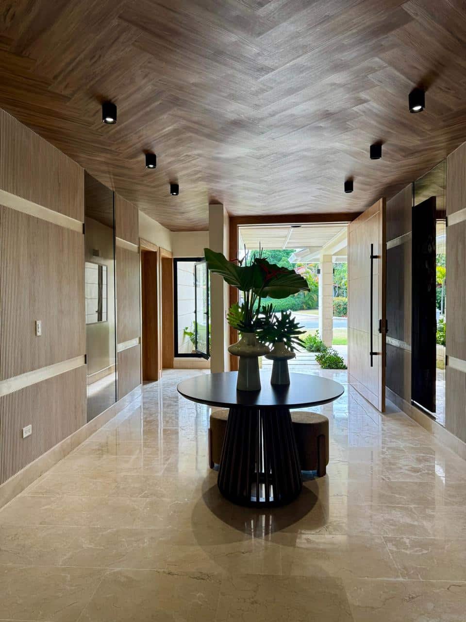 Vestíbulo con techo de madera, paredes decoradas con paneles y puertas de madera, mesa redonda con jarrones y plantas frente a la entrada con vista al exterior y áreas verdes.