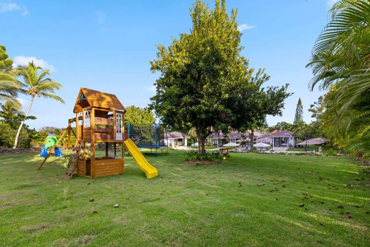 Jardín con césped, columpio y resbaladilla de madera, árbol grande, casas en el fondo, paraguas y sillas de jardín.