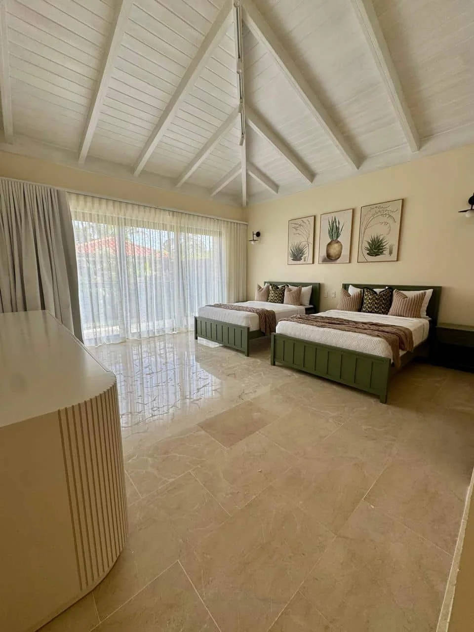 Habitación con techos altos de madera pintados de blanco, dos camas de madera verde con ropa de cama clara, cuadros decorativos con plantas en la pared, cortinas blancas que dejan ver el exterior.