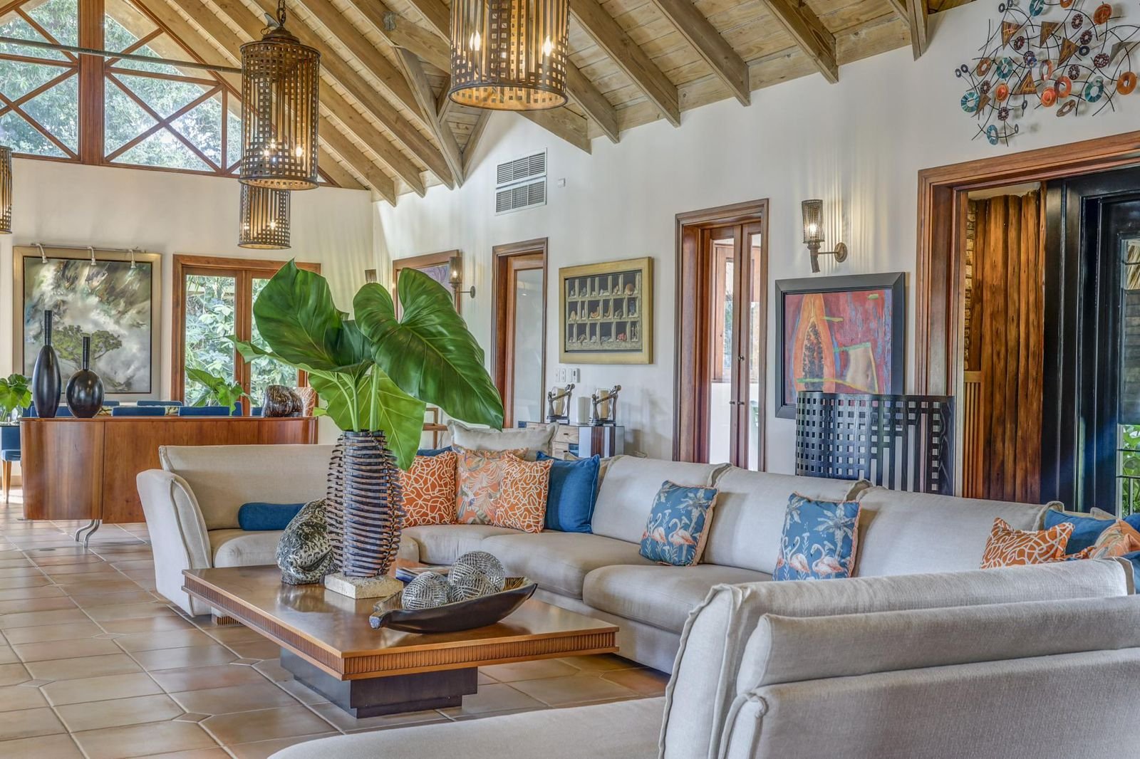 Salón con sofá beige adornado con cojines de colores y patrón floral, mesa de madera con decoración de plantas y bolas, paredes blancas con cuadros y esculturas, ventanas de madera, techo de madera con vigas expuestas, iluminación cálida.