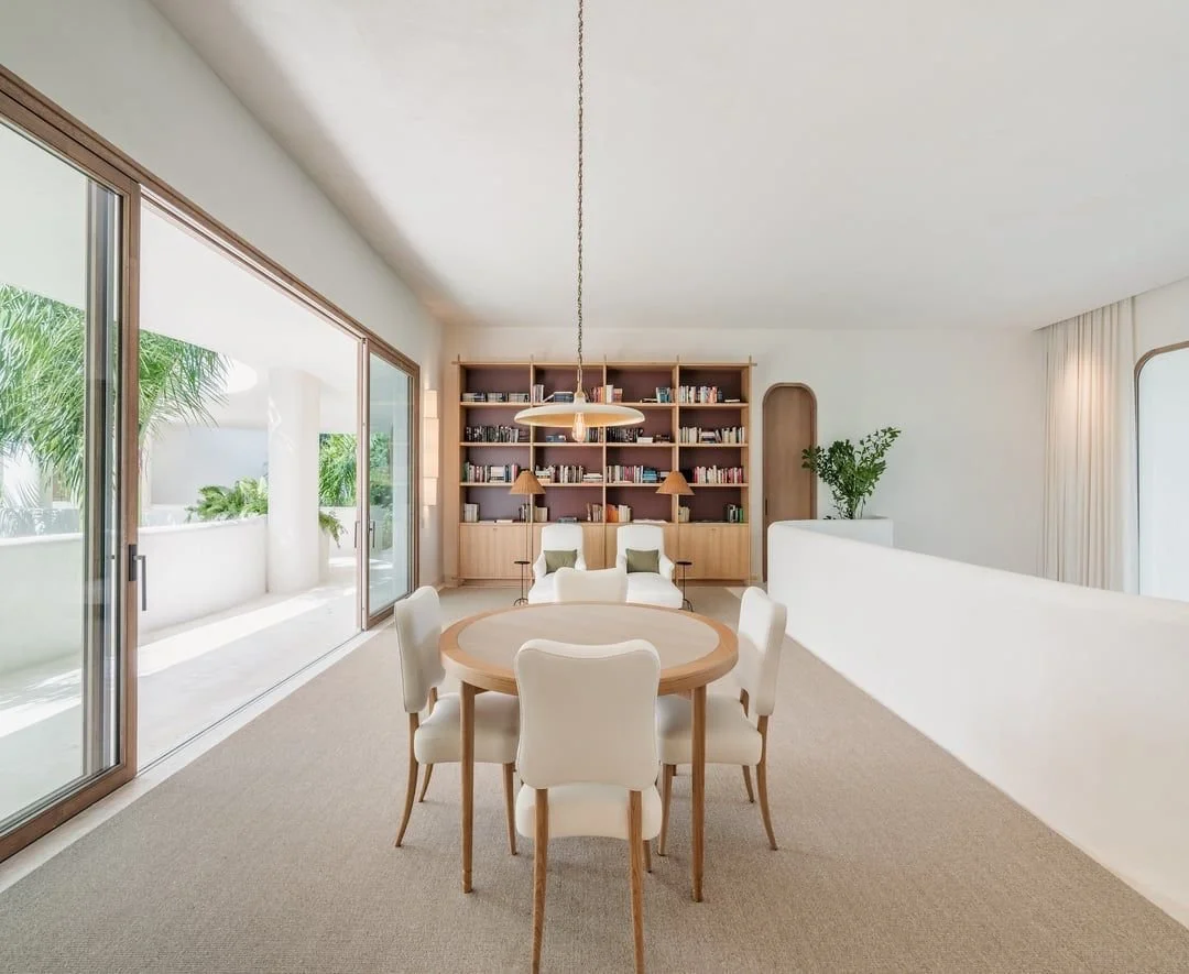 Sala de estar con mesa redonda, sillas blancas, estantería llena de libros, ventanas grandes con vista a un balcón con plantas tropicales.