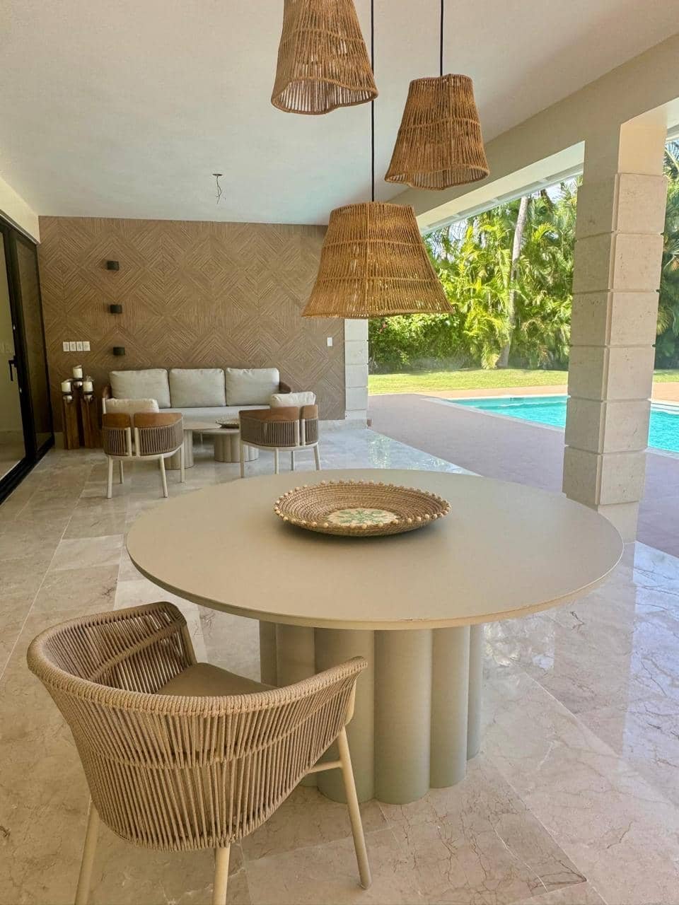 Área de terraza con mesa redonda, silla de mimbre, sillón blanco y lámparas de borde de mimbre, vista a la piscina y vegetación tropical.