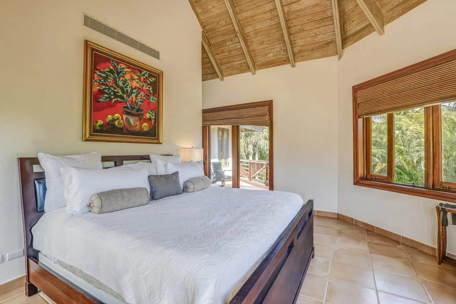 Habitación con cama grande, pintura de un jarrón con plantas y frutas, ventanas con vistas a la naturaleza, techo de madera