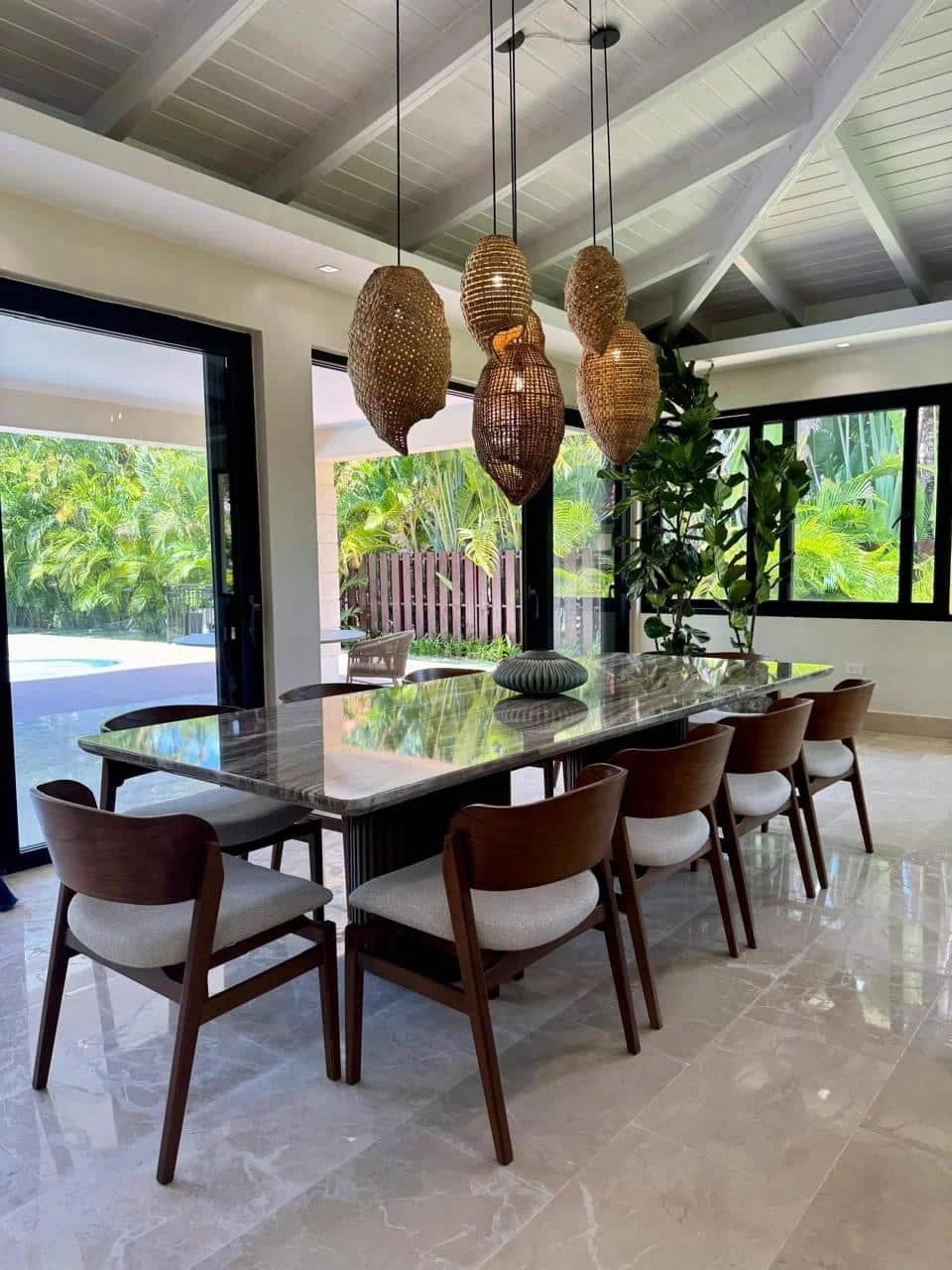 Comedor interior con mesa de mármol, ocho sillas de madera con cojines claros, lámparas colgantes de mimbre, ventanas grandes con vista a un jardín con plantas verdes.