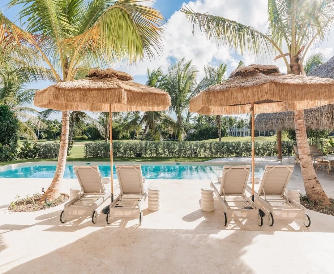Sillas de descanso junto a una piscina con sombrillas de paja, palmeras y vegetación tropical alrededor.