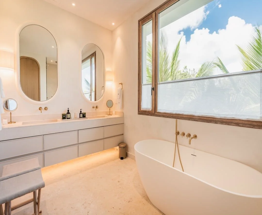 Baño moderno con tina freestanding, dos espejos ovalados sobre lavabo, ventana grande con vista a plantas tropicales y cielo azul.