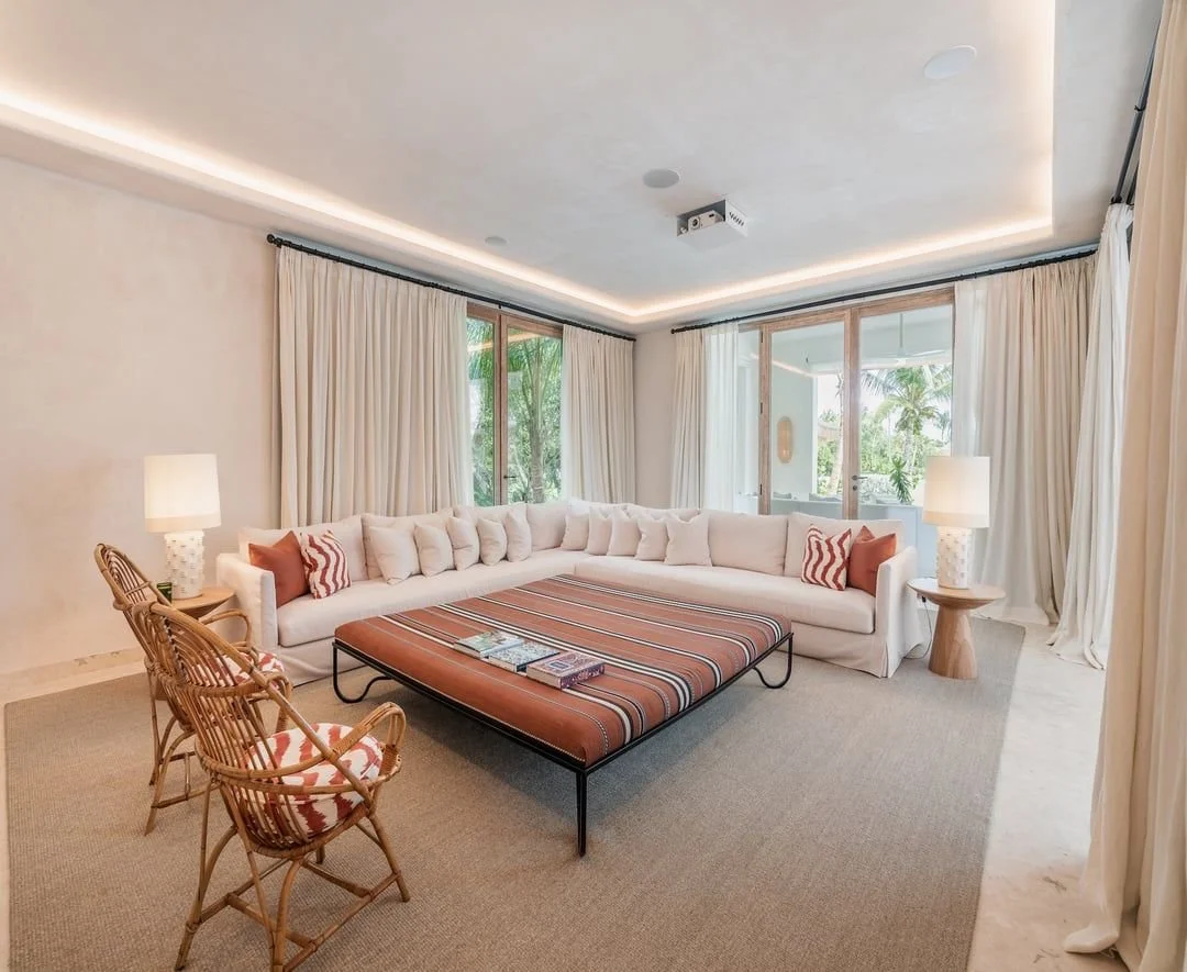 Sala de estar con sillón grande en forma de L, mesa de centro con cojines y sillas de mimbre, cortinas blancas y vista a un balcón con plantas.