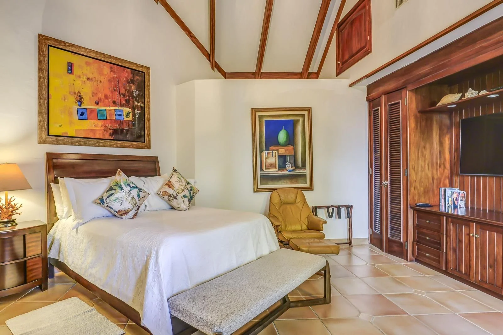 Habitación con cama de matrimonio, sillón de cuero, armario grande de madera, cuadros decorativos, suelo de baldosas de barro, lámpara junto a la cama.