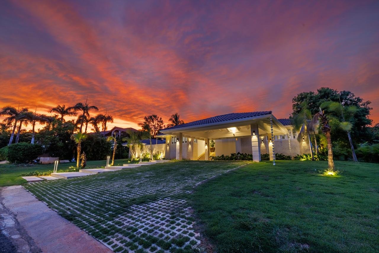 Casa moderna con iluminación exterior y palmeras, vista de atardecer con cielo nublado en tonos anaranjados y púrpuras.