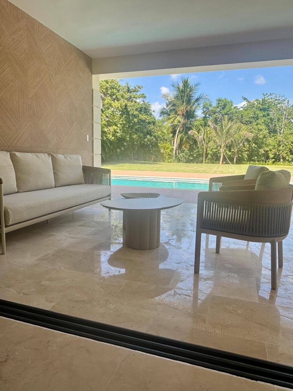 Espacio de sala con sillones beige y una mesa redonda, vista al patio con piscina y árboles en un día soleado.