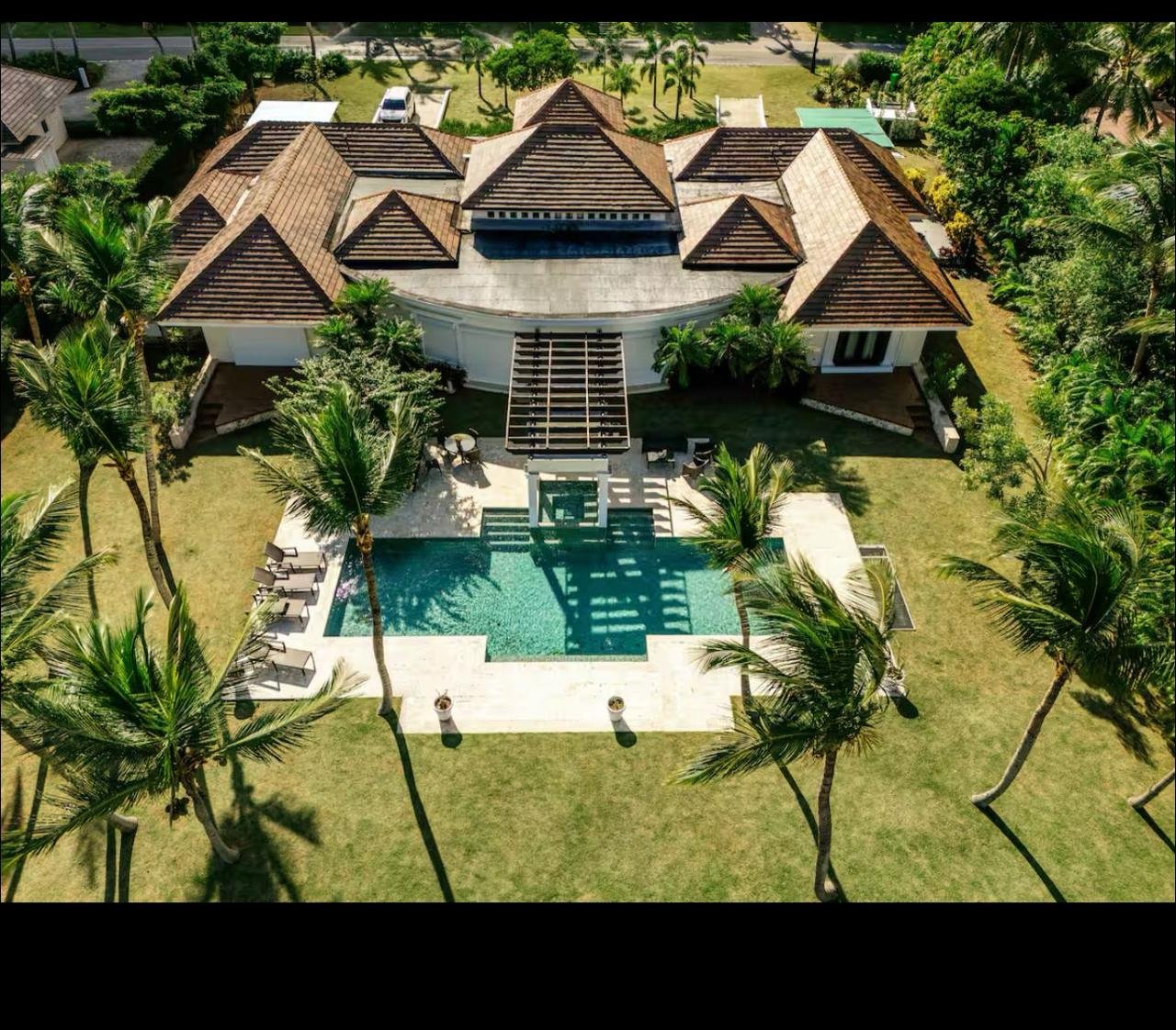 Vista aérea de una casa grande con tejados de teja en medio de un jardín con palmeras y alberca.