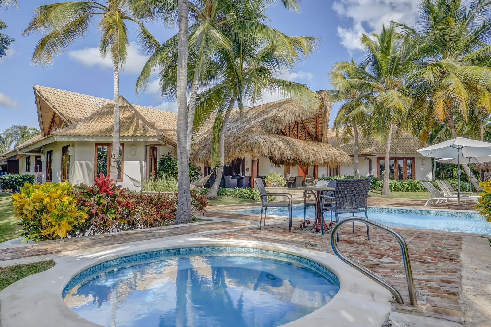 Vista de una casa en la playa con piscina y área de descanso con sombrillas, rodeada de palmeras y vegetación tropical, en un día soleado con cielo azul.