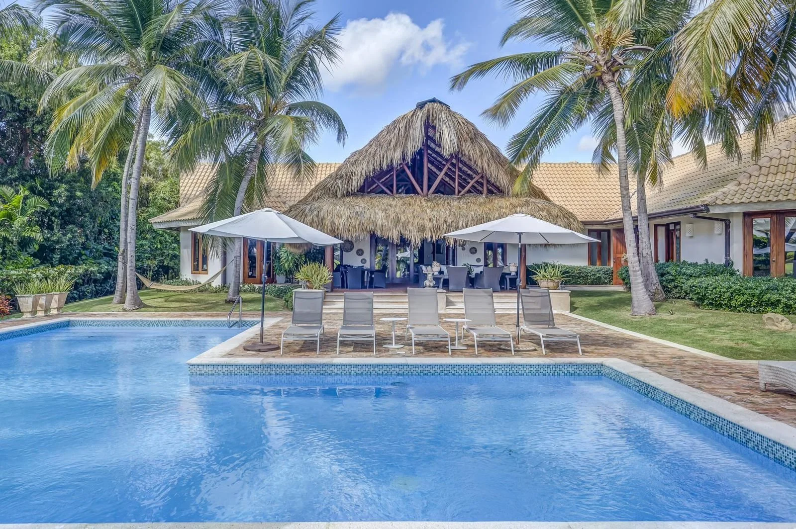 Casa de playa con piscina, sombrillas, sillas de playa y palmeras en un entorno tropical
