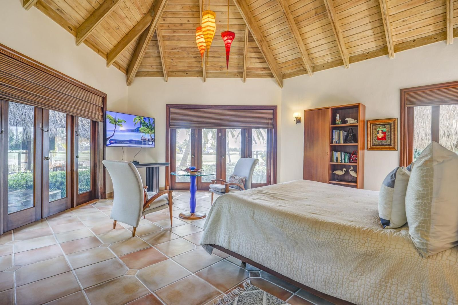 Habitación con cama, sillas, televisión, ventanas con vista a palmeras y decoraciones tropicales.