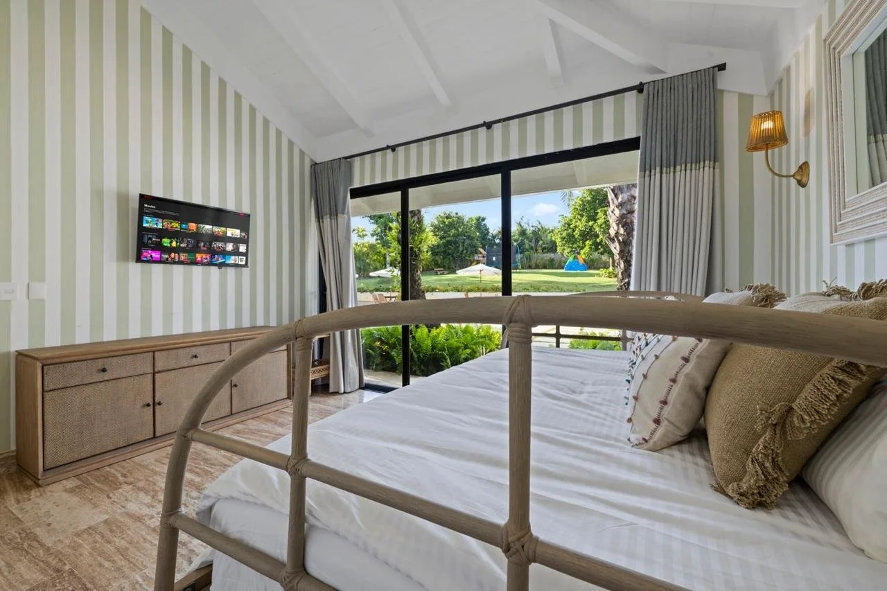 Habitación con cama, vista al jardín exterior, cortinas, televisor en la pared y paredes con rayas verdes y blancas.