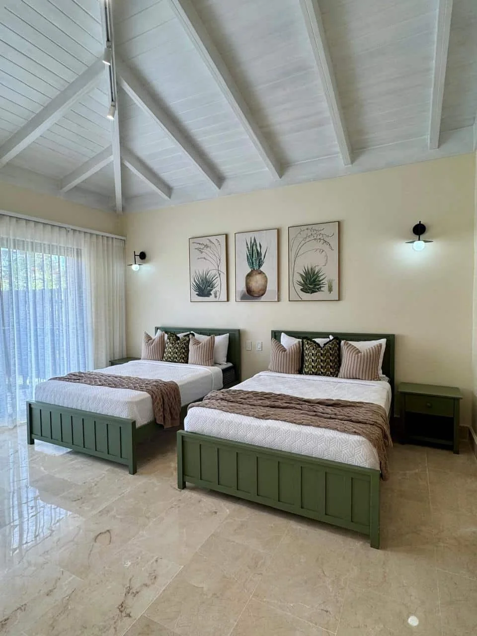 Habitación con dos camas dobles con cabeceras verdes, ropa de cama blanca y cojines decorativos, arte de plantas en la pared, cortinas transparentes y lámparas de pared.