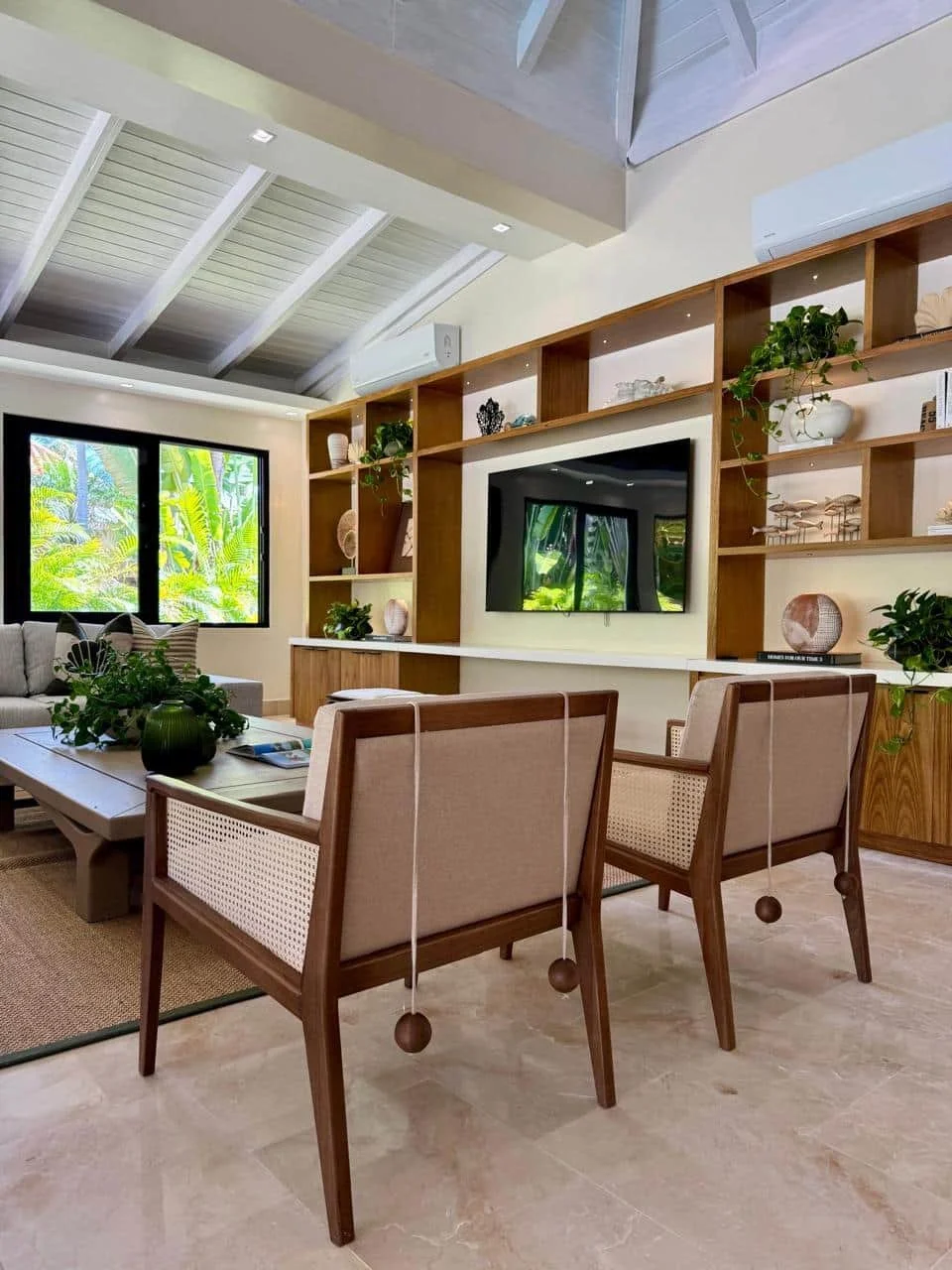 Sala de estar con sillones de madera y tela, mesa centro con plantas y libros, paredes con ventanas y estanterías con decoraciones y plantas, televisión, paredes blancas y techo de madera y vidrio.