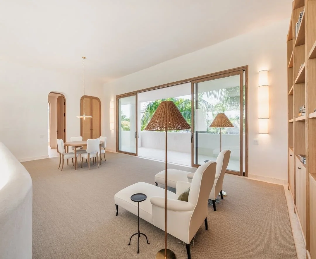 Sala de estar con sillones blancos, lámparas de mimbre, mesa auxiliar negra, librero de madera, puerta corredera que da a un balcón con vista a plantas tropicales.