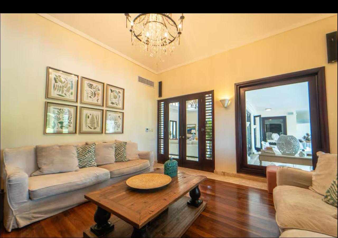 Sala de estar con dos sofás beige, mesa de madera con decoración, cuadros con plantas en la pared, lámpara de araña en el techo y ventanas con contraventanas de madera.