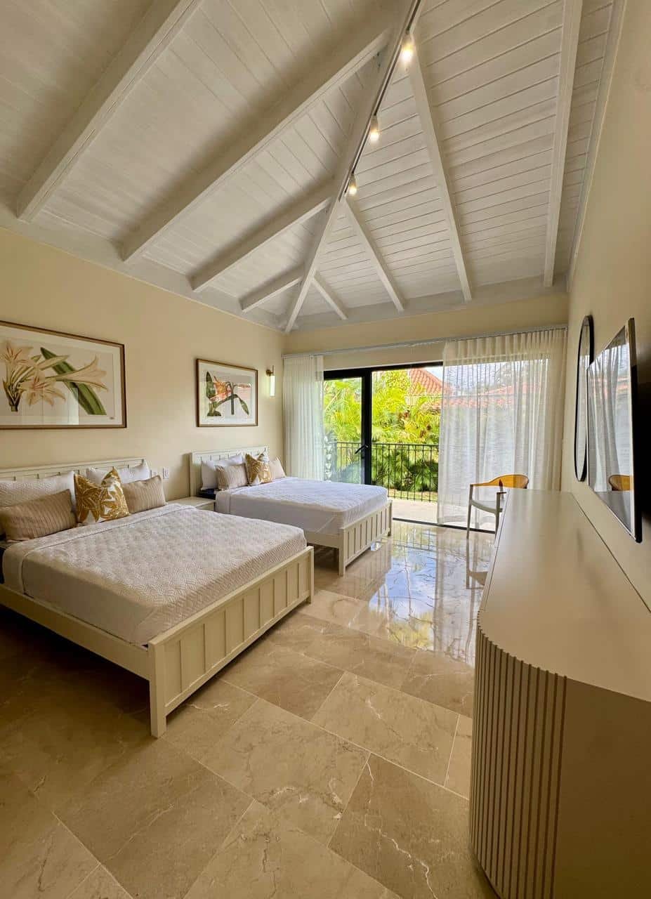 Habitación con techo alto de vigas blancas, dos camas con cabecera blanca y colchas blancas, varias almohadas beige y doradas, arte decorativo en las paredes y vista a un balcón con vegetación tropical.