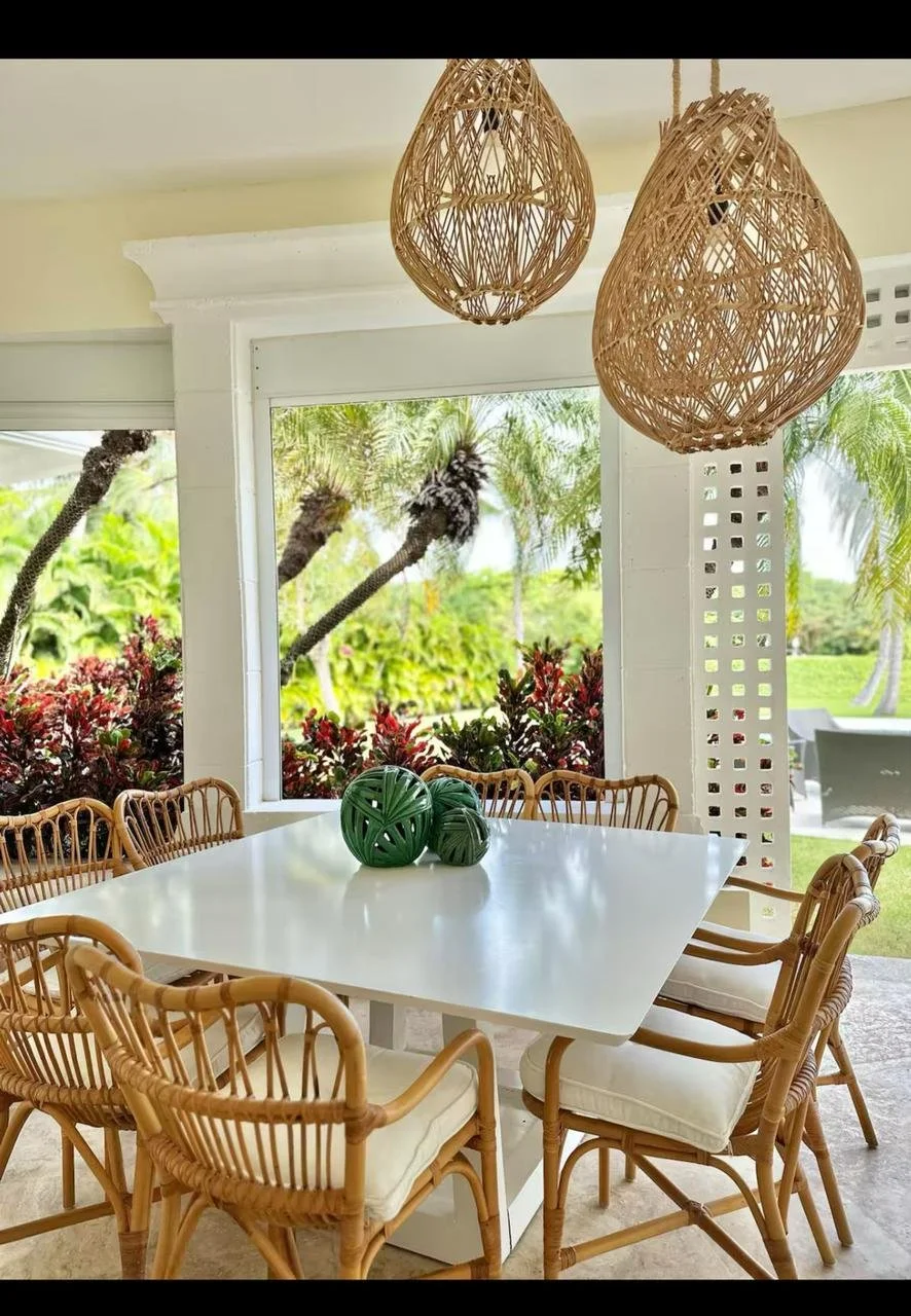 Mesa blanca con tres decorativos verdes en el centro en un comedor con sillas de mimbre, vista de un jardín con palmeras y follaje desde una ventana grande y luminosa.