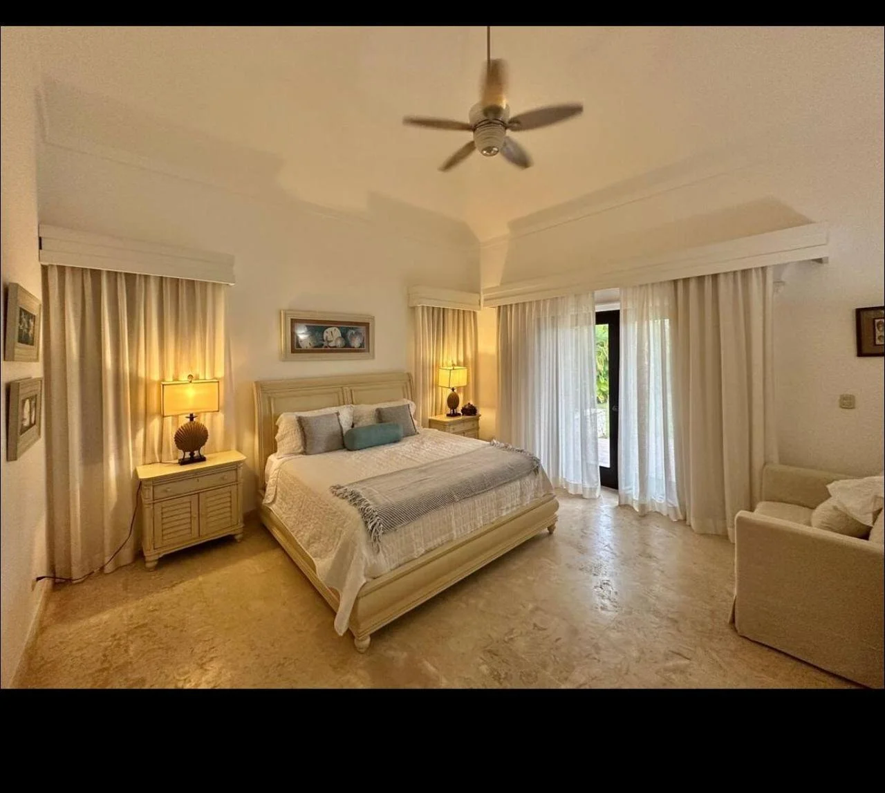 Habitación con cama grande, cortinas blancas, lámparas de mesa, sillón beige y puertas con vista al exterior