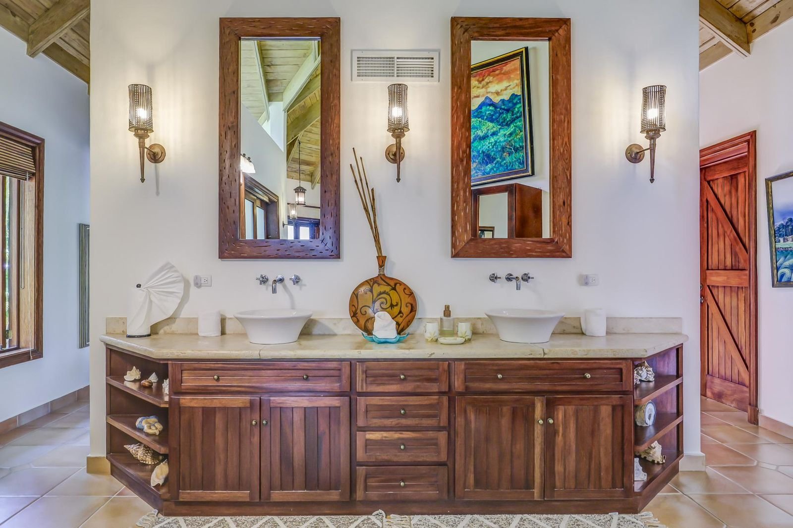 Un baño con doble lavabo, dos espejos grandes con marcos de madera, lámparas de pared en ambos lados, y decoración en la repisa central con un jarrón de cerámica, velas y señas decorativas. Ventanas de madera y una puerta de madera a la derecha.