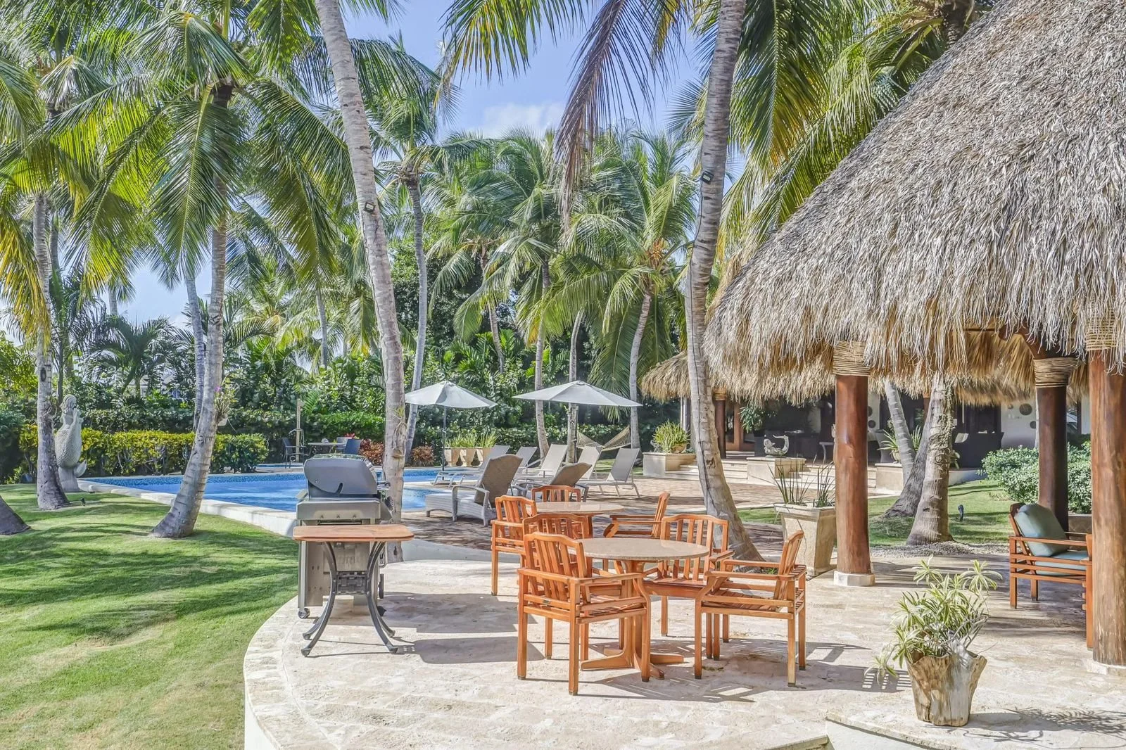 Áreas al aire libre con piscina, sombrillas, muebles de patio, palmeras y estructura de madera con techo de paja en un ambiente tropical
