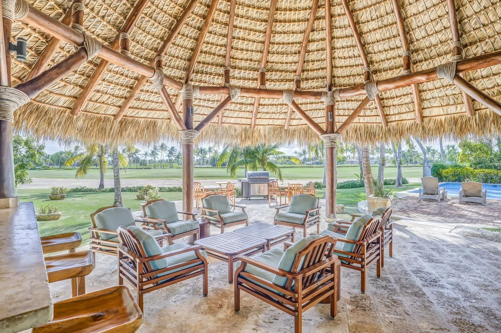 Área de descanso al aire libre con muebles de madera y cojines en una estructura de techo de palma, con vista a un campo de golf con palmeras y una piscina en el fondo.