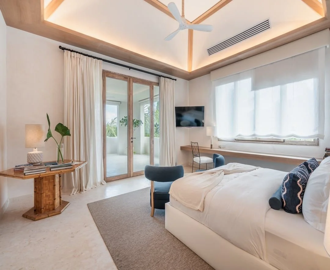 Habitación con cama grande, sillones azules, cortinas blancas, televisor, lámparas, balcón con plantas y puertas de vidrio, techo de madera con ventilador.