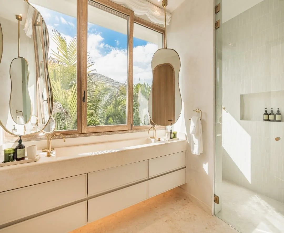 Baño con doble lavabo, espejo y ventana que muestra plantas y cielo azul.