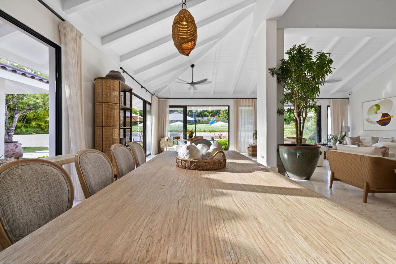 Interior de una sala de comedor luminosa con mesa de madera y sillas tapizadas, plantas en macetas grandes y ventanas con cortinas claras que dejan ver un espacio exterior con árboles y sombrillas.