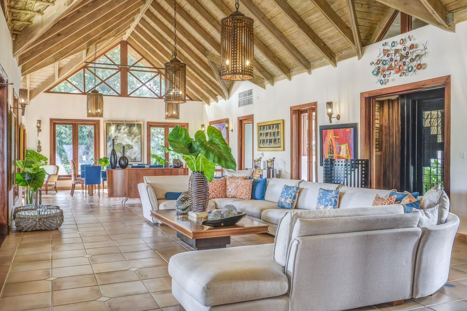 Sala con sofá blanco y cojines coloridos, paredes blancas, ventanas grandes, techo de madera con vigas, lámparas colgantes de estilo rústico, decoraciones en las paredes, plantas verdes, mesa de centro de madera.