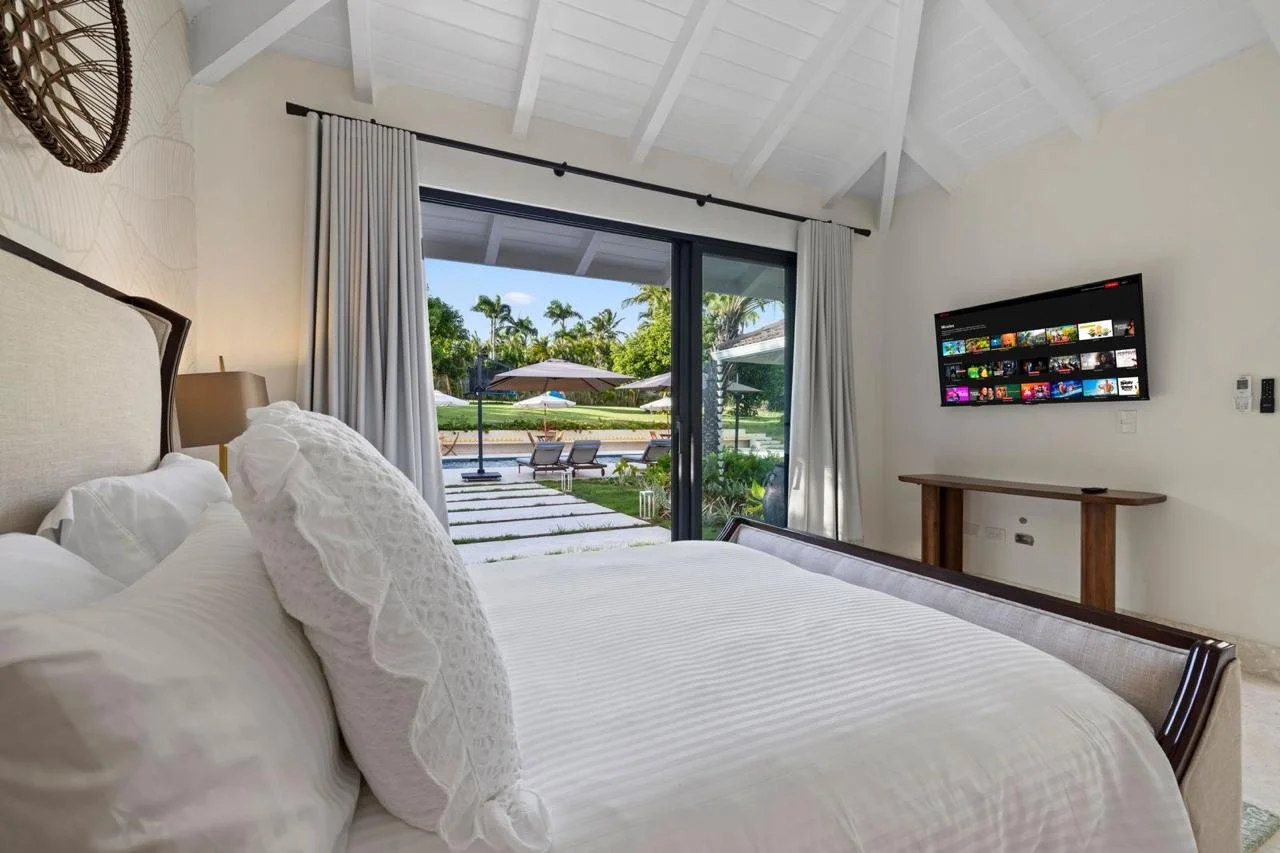 Habitación con cama blanca, vista al jardín con sombrillas y sillas, televisor en la pared y cortinas blancas.