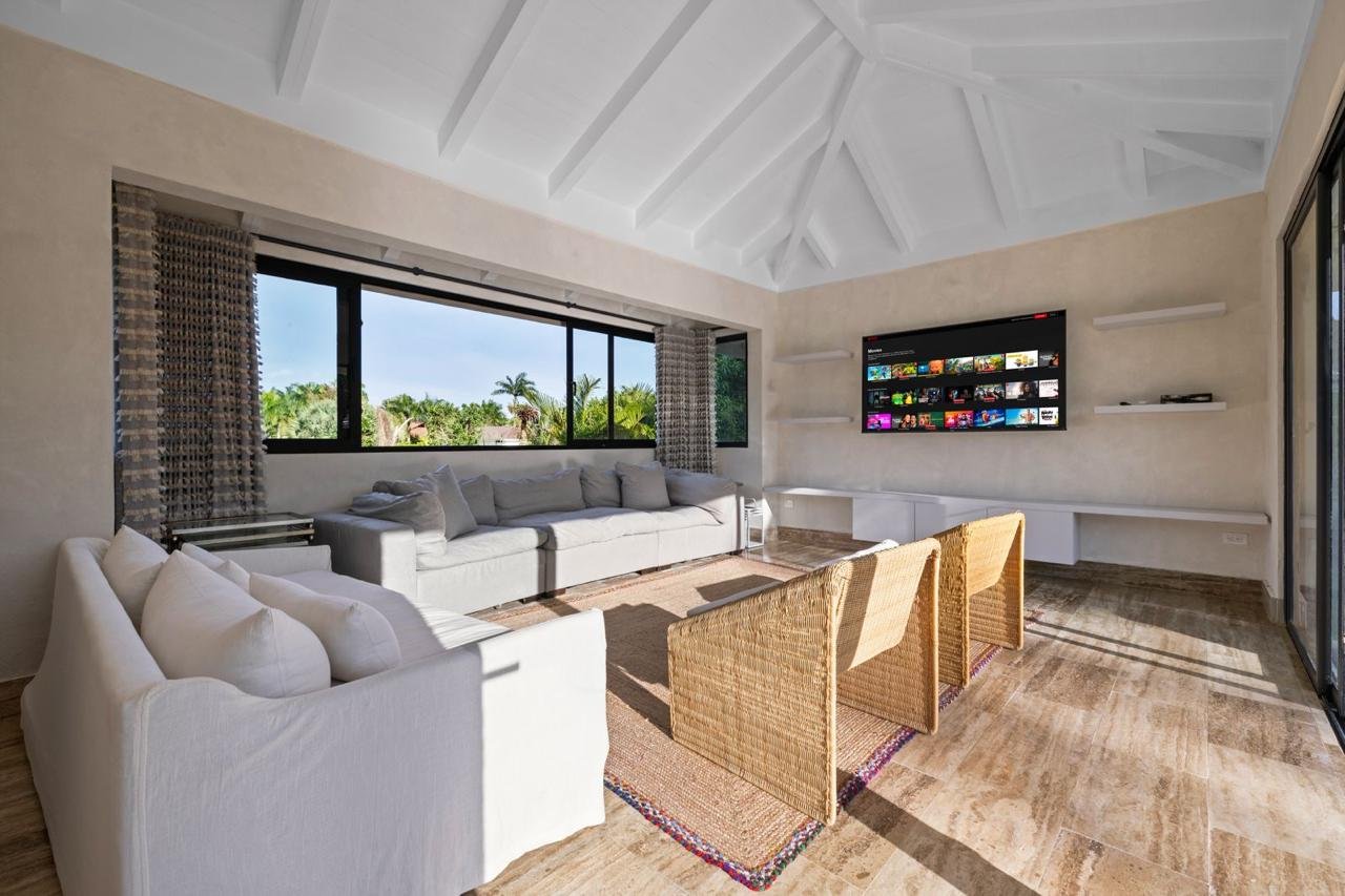 Sala de estar con sillones blancos, una pantalla plana en la pared y ventanas grandes que muestran plantas y cielo azul.