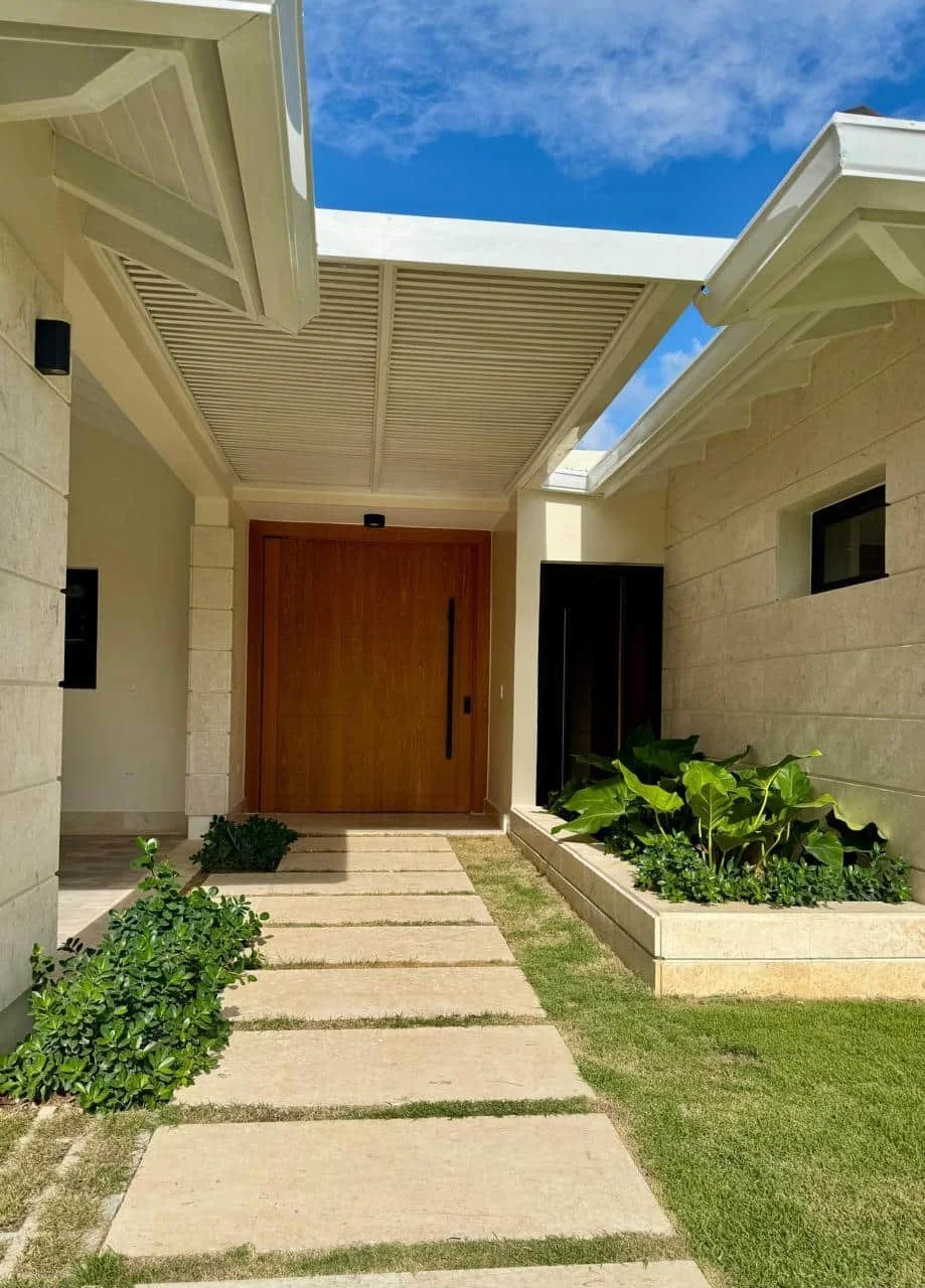 Entrada principal de una casa moderna con camino de loseta, plantas verdes a los lados y puerta de madera, bajo cielo azul.