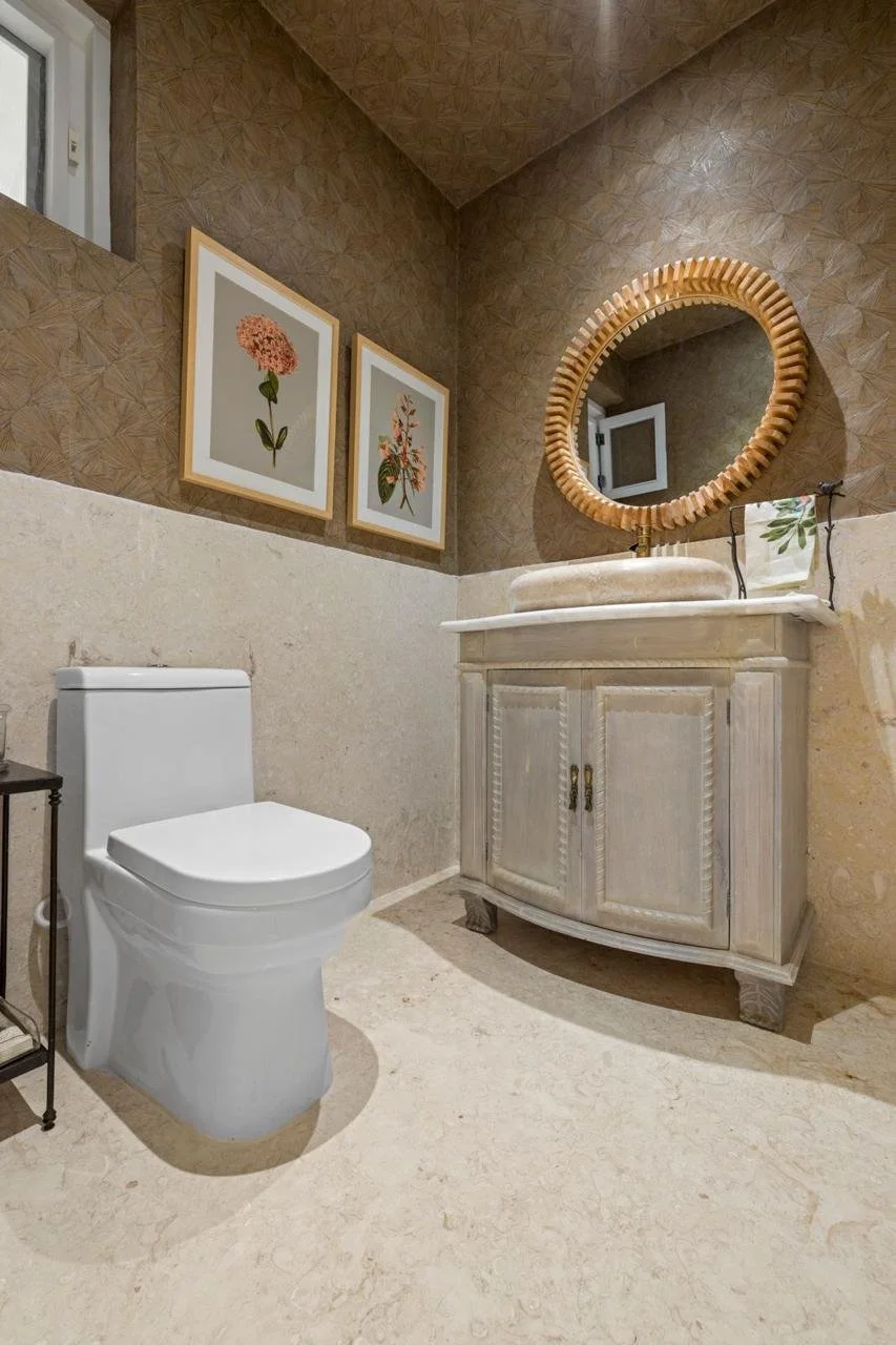 Baño con inodoro blanco, lavabo con mueble de madera lacada en blanco, espejo redondo de marco de madera, pared decorada con papel tapiz de patrón geométrico en tonos marrón, y dos cuadros con flores en la pared.
