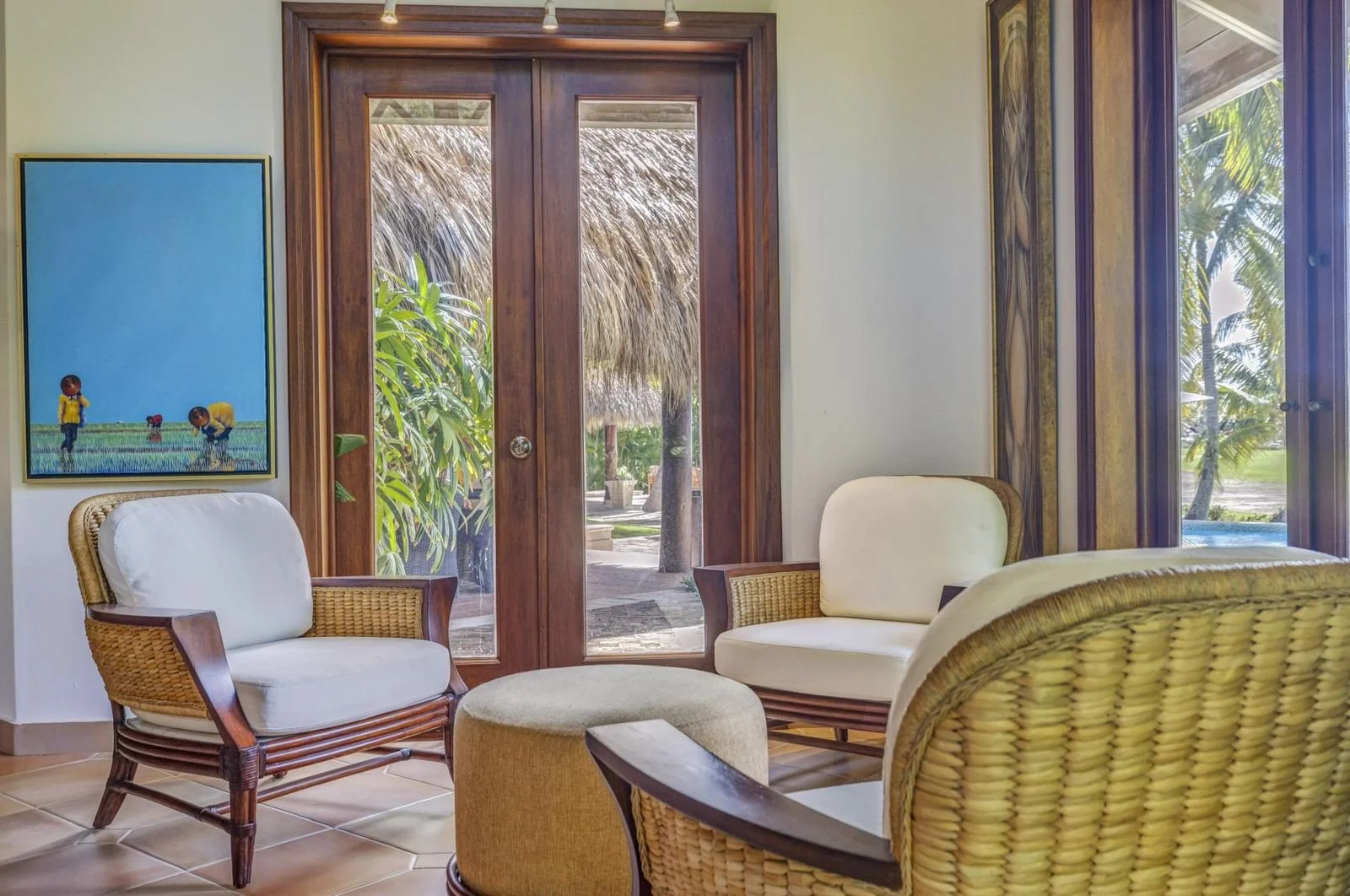 Sala de estar con sillones de mimbre y cojines blancos, puertas de madera con vista a un jardín tropical con palmeras y techo de palma.